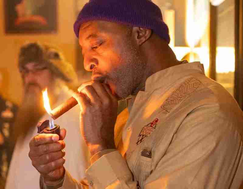 A man in a purple beanie and a tan guayabera shirt uses a torch lighter to light a premium cigar inside a warm, dimly lit lounge during a holiday party.