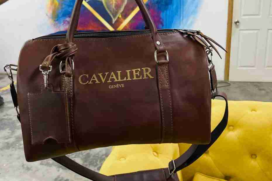 A premium brown leather Cavalier Genève duffel bag with gold branding text held up above a yellow tufted couch in a lounge or studio setting.