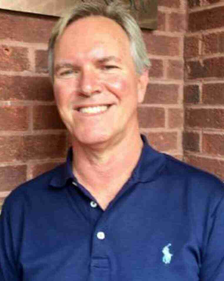 F&M Cigars Staff 1 A close-up portrait of Brad Williams smiling in a navy blue Polo Ralph Lauren shirt, standing outdoors in front of a textured red brick wall.