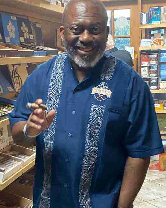 F&M Cigars Staff 9 A smiling, bearded man holding a premium cigar stands inside a well-stocked humidor shop. He is wearing a navy blue F&M Cigars embroidered guayabera shirt, surrounded by wooden shelves displaying various cigar boxes.