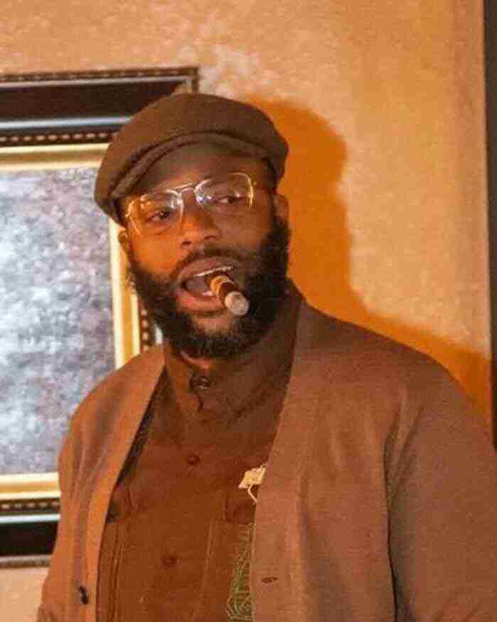 F&M Cigars Staff 7 A bearded man named Kamorris Johnson wearing clear aviator glasses, a brown flat cap, and a matching brown cardigan enjoys a premium cigar. He is captured mid-conversation under warm indoor lighting in front of a textured wall and a framed picture.