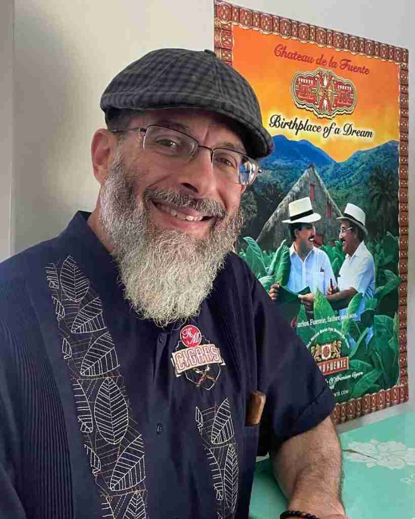 F&M Cigars Staff 3 A smiling bearded man in a dark blue F&M Cigars embroidered guayabera shirt and flat cap poses in front of a colorful Arturo Fuente "Chateau de la Fuente" poster.