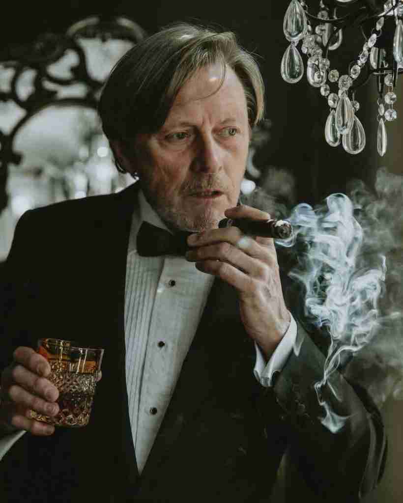 F&M Cigars Staff 5 A man in a formal black tuxedo and bow tie holds a cut-glass tumbler of whiskey while exhaling thick white smoke from a premium cigar. He is set against a moody background featuring a crystal chandelier.