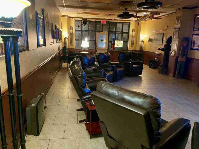 membership-lounge - 1 Rows of comfortable black leather recliner chairs inside the spacious, warmly lit lounge area at F&M Cigars, featuring tiled floors, wood-paneled walls, and classic decor.