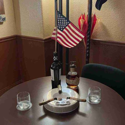 membership-lounge - 5 A table setup at F&M Cigars featuring a ceramic ashtray with two resting cigars, rocks glasses on coasters, a bottle of premium bourbon, and a small American flag.