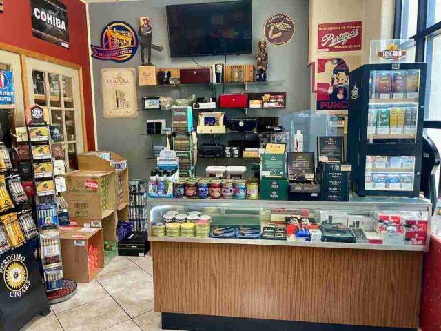 walk-in-humidor - 10 The main retail checkout counter at F&M Cigars, featuring a glass display case with premium cutters and lighters, wall shelving stocked with accessories, and advocacy posters.