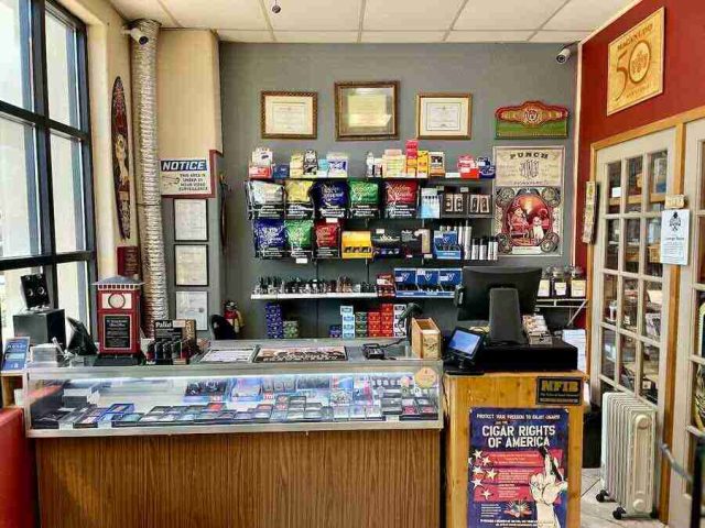 walk-in-humidor - 12 The main retail checkout counter at F&M Cigars, featuring a glass display case with premium cutters and lighters, wall shelving stocked with accessories, and advocacy posters.