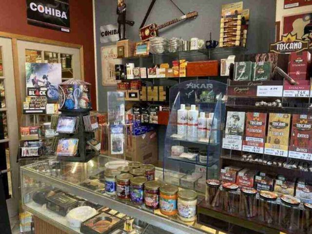 walk-in-humidor - 15 The main retail checkout counter at F&M Cigars, featuring a glass display case with premium cutters and lighters, wall shelving stocked with accessories, and advocacy posters.