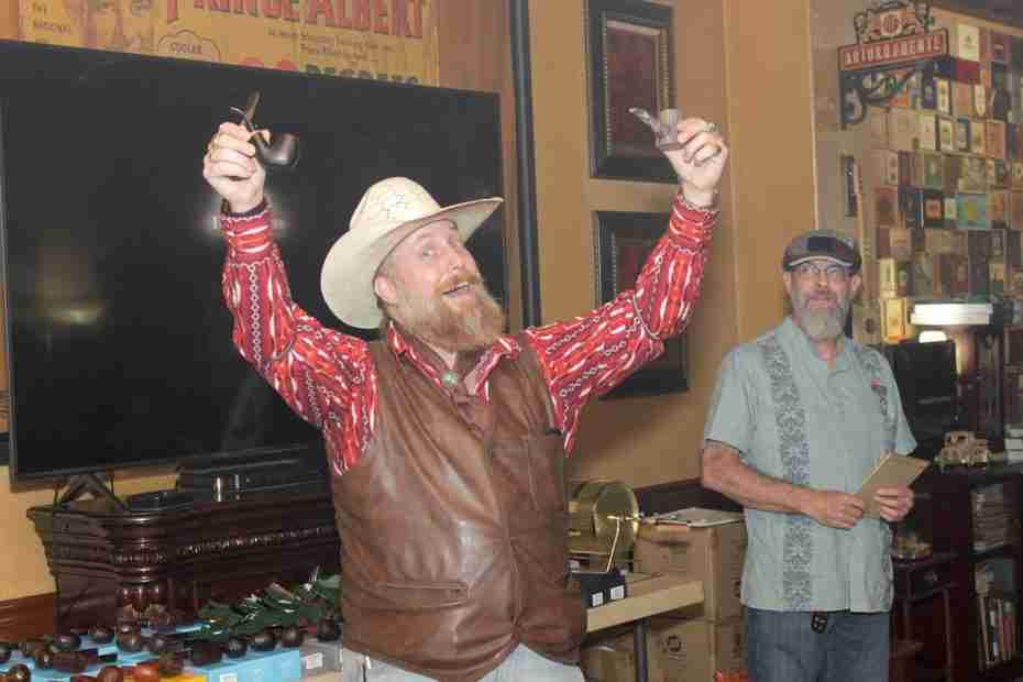 A cheerful, bearded man in a cowboy hat, red pattern-embroidered shirt, and brown leather vest triumphantly holds up two smoking pipes in front of a flat-screen TV, while a man in a grey shirt and cap stands alongside him in a lounge decorated with cigar boxes and vintage tobacco art.